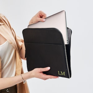 Könnte beinhalten: Eine schwarze Laptoptasche mit Reißverschluss und einem grünen Monogramm "LM" auf der Vorderseite. Die Tasche wird von einer Person gehalten.