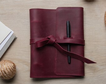 May include: A burgundy leather journal with a matching tie closure and a black pen inserted in the spine. The journal is closed and resting on a light wood surface, with decorative spheres and a woven basket in the background.