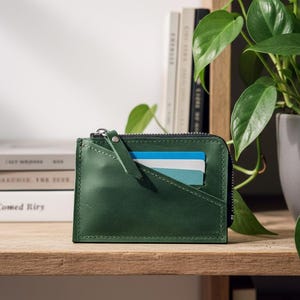 Może przedstawiać: Ciemnozielony skórzany portfel z zapięciem na zamek błyskawiczny i przegródką na karty. Portfel jest prostokątny i ma mały skórzany pasek przymocowany do zamka. Kilka kart kredytowych jest widocznych w przegródce.