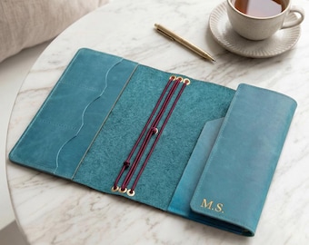 May include: A teal leather notebook cover featuring three vertical burgundy elastic bands, gold-toned metal eyelets, and interior pockets with scalloped edges. The right flap is embossed with the gold-colored initials M.S. in a serif font.