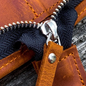 May include: Close-up of a brown leather zipper pouch with a silver zipper pull. The zipper is open, revealing the black fabric lining. The pouch has a brown leather strap with a silver rivet.