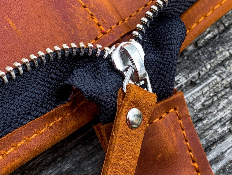 May include: Close-up of a brown leather zipper pouch with a silver zipper pull. The zipper is open, revealing the black fabric lining. The pouch has a brown leather strap with a silver rivet.