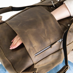May include: A brown leather backpack with a large front pocket and a zippered pocket on the side. The backpack has a distressed look and is made of high-quality leather.