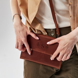 May include: A brown leather crossbody bag with a silver clasp and a monogrammed initial "S" surrounded by leaves.