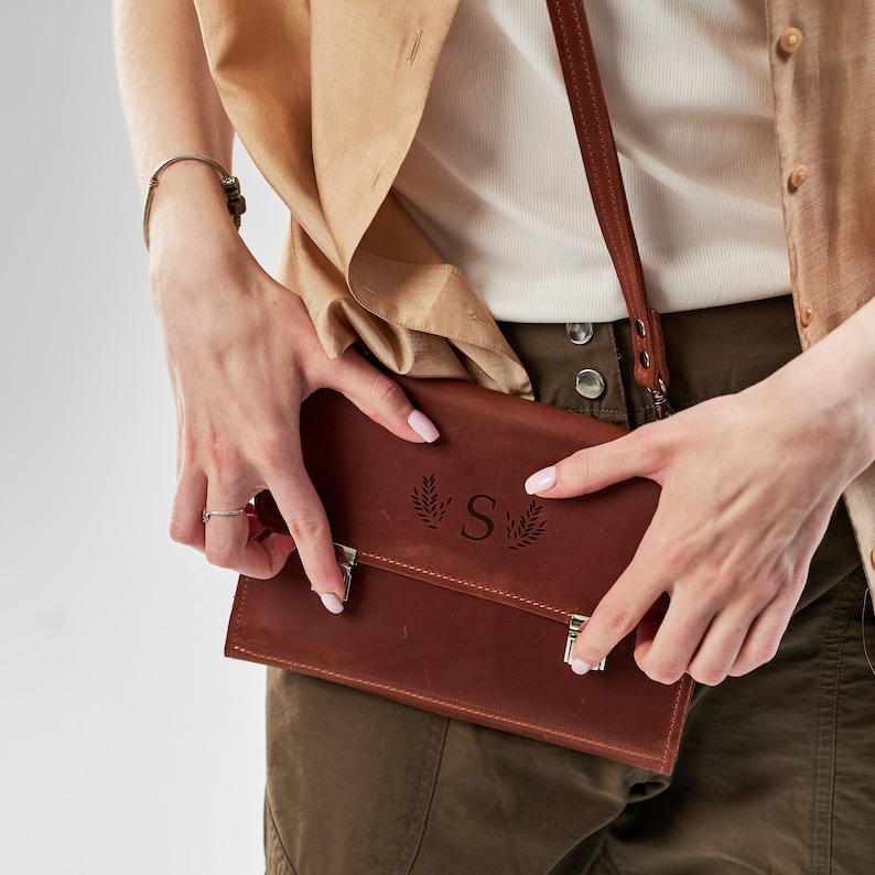May include: A brown leather crossbody bag with a silver clasp and a monogrammed initial "S" surrounded by leaves.