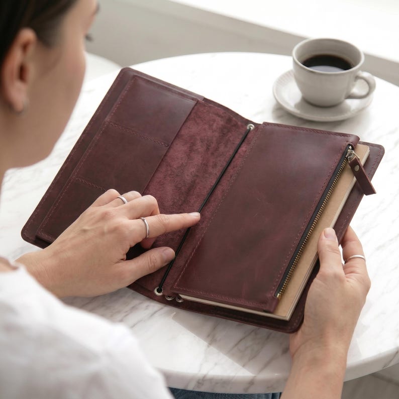 May include: A dark burgundy leather journal cover featuring an open design with internal pockets, a zippered compartment, and an elastic band closure. The interior displays a smooth, matte finish with visible stitching and a notebook insert.
