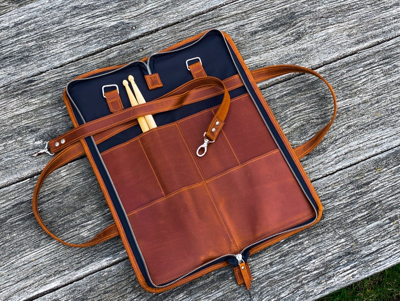 May include: A brown leather drum stick bag with a black canvas lining. The bag has multiple pockets and a zipper closure. The bag is open and two wooden drum sticks are inside.