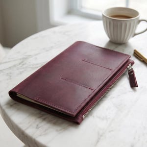 May include: A deep burgundy leather notebook cover featuring a side zipper closure with a matching leather pull tab and two horizontal stitched slots on the front cover. The rectangular cover is closed and rests flat on a surface.