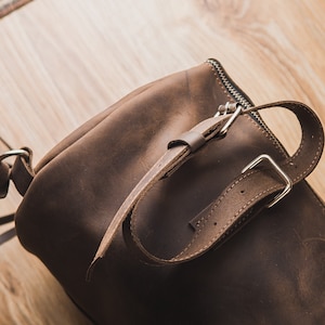 Puede incluir: Un bolso de cuero marr&oacute;n con una hebilla plateada y un cierre de cremallera. El bolso tiene una correa que se puede ajustar para adaptarse a diferentes tama&ntilde;os.