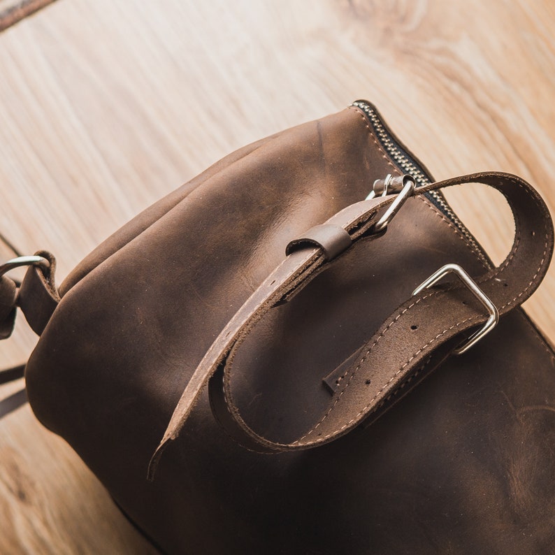 Puede incluir: Un bolso de cuero marr&oacute;n con una hebilla plateada y un cierre de cremallera. El bolso tiene una correa que se puede ajustar para adaptarse a diferentes tama&ntilde;os.