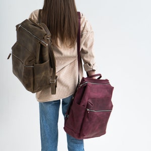 May include: Two leather backpacks, one brown and one burgundy, worn by a person. The brown backpack has a zippered pocket on the front. The burgundy backpack has a zippered pocket on the front and a zippered pocket on the side.