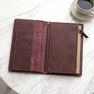 May include: A dark brown leather notebook cover featuring an open design with multiple interior pockets on the left side, a central elastic cord for securing inserts, and a zippered pouch on the right side with a matching leather pull tab.