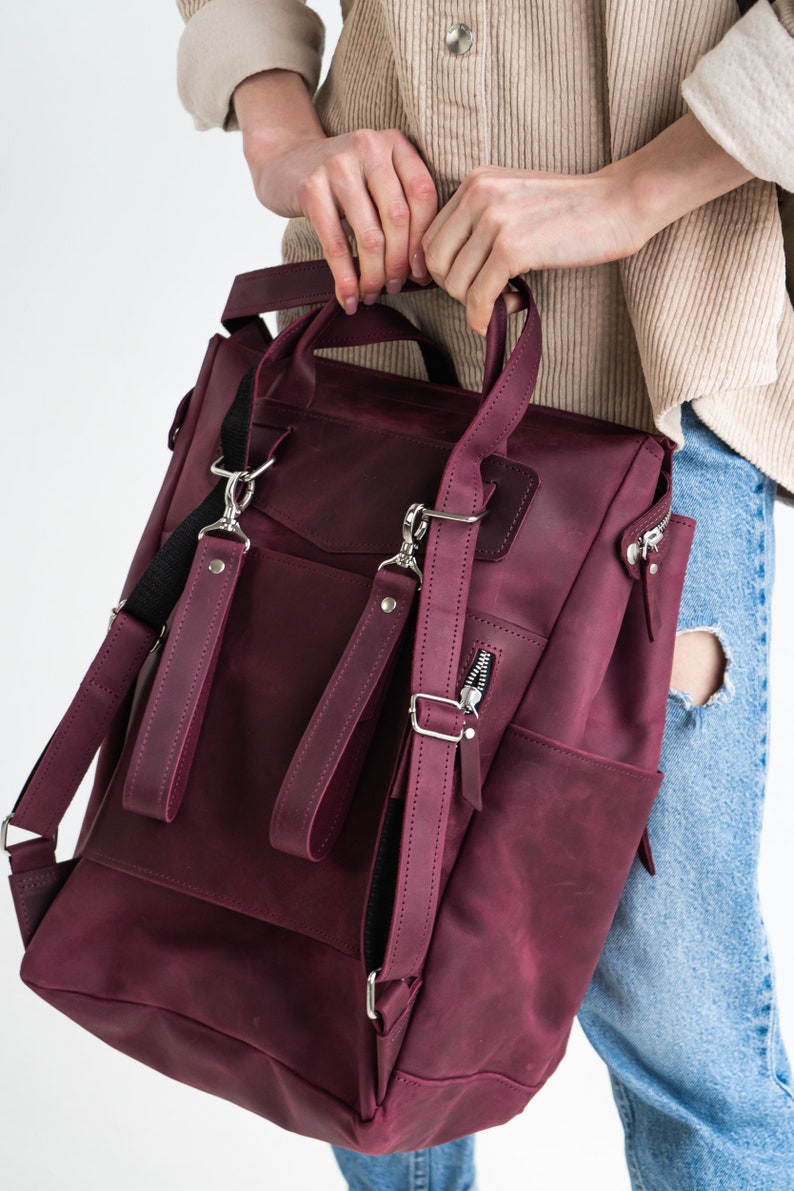 May include: A burgundy leather backpack with adjustable straps and multiple pockets. The backpack has a zippered front pocket and a flap closure.