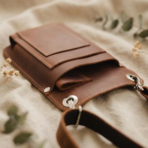 K&ouml;nnte beinhalten: Eine braune Leder-Umh&auml;ngetasche mit Klappenverschluss. Die Tasche hat eine kleine Vordertasche und einen langen, verstellbaren Riemen. Die Tasche ist aus strukturiertem Leder gefertigt und hat silberfarbene Beschl&auml;ge.