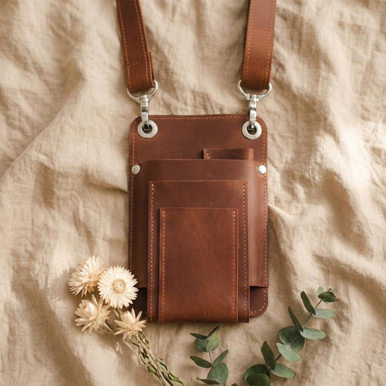 K&ouml;nnte beinhalten: Eine braune Leder-Umh&auml;ngetasche mit mehreren Taschen und einem Schultergurt. Die Tasche hat silberfarbene Hardware und ist rechteckig geformt. Die Tasche wird auf einem beigefarbenen Stoffhintergrund mit Trockenblumen und Gr&uuml;nzeug gezeigt.
