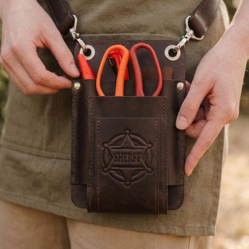 K&ouml;nnte beinhalten: Ein brauner Leder-Werkzeugg&uuml;rtel mit einem Sheriff-Abzeichen-Design. Der G&uuml;rtel hat mehrere Taschen mit orangefarbenen und roten Scheren und einem roten Stift. Der G&uuml;rtel wird von einem braunen Riemen mit silbernen Metallverschl&uuml;ssen getragen.