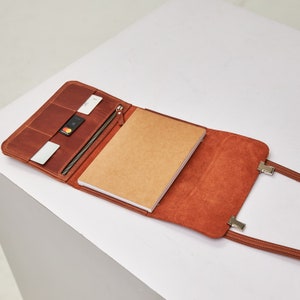 May include: A brown leather portfolio with a zipper closure and a notebook inside. The portfolio has a strap with a metal clasp.