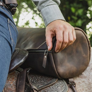 Puede incluir: Una bolsa de lona de cuero marr&oacute;n con cierre de cremallera y una correa de cuero. La bolsa est&aacute; siendo sostenida por una mano y est&aacute; sentada sobre una silla de montar.