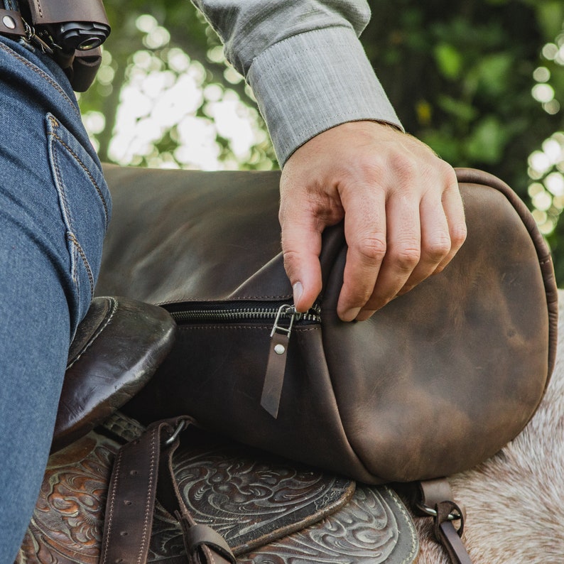 Puede incluir: Una bolsa de lona de cuero marr&oacute;n con cierre de cremallera y una correa de cuero. La bolsa est&aacute; siendo sostenida por una mano y est&aacute; sentada sobre una silla de montar.