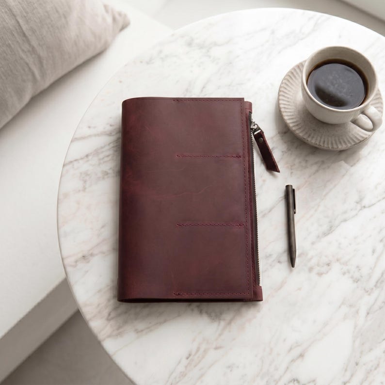 May include: A dark burgundy leather notebook cover featuring a vertical side zipper closure, a matching leather zipper pull, and three horizontal stitched lines on the front cover, accompanied by a slim dark metal pen.