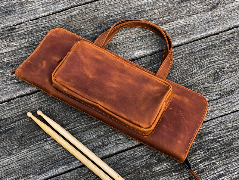 May include: A brown leather drum stick bag with a smaller brown leather pouch attached. The bag has a handle and a zipper closure. Two wooden drum sticks are lying on a wooden surface.