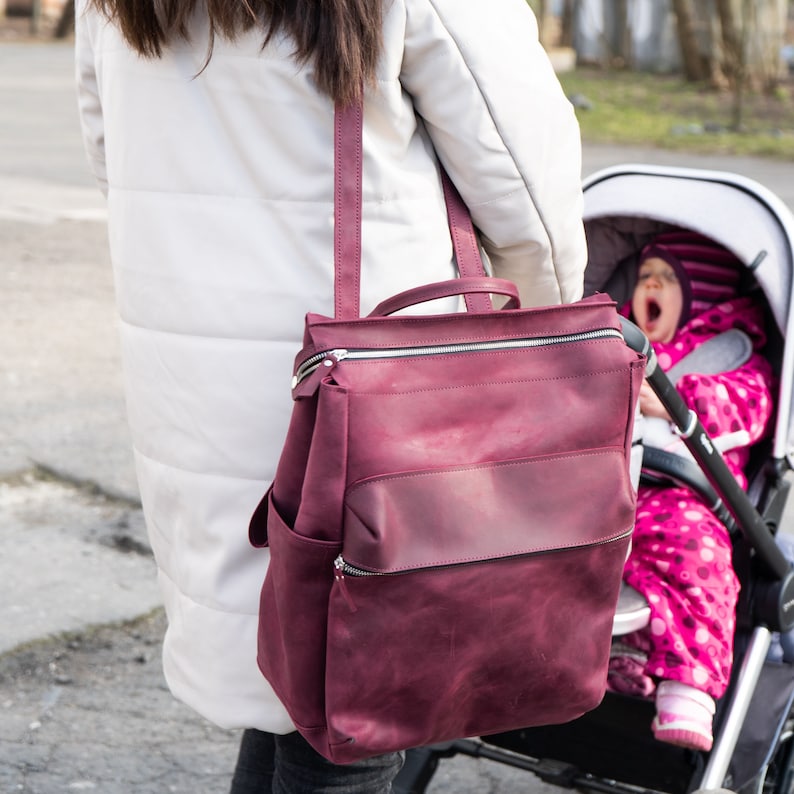 May include: A woman wearing a white coat is pushing a stroller with a baby inside. The woman is carrying a large burgundy leather backpack with multiple zippered compartments.