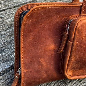 May include: A brown leather zippered pouch with a smaller zippered pouch attached. The main pouch has a flap that folds over and closes with a zipper. The smaller pouch is on the right side of the main pouch.