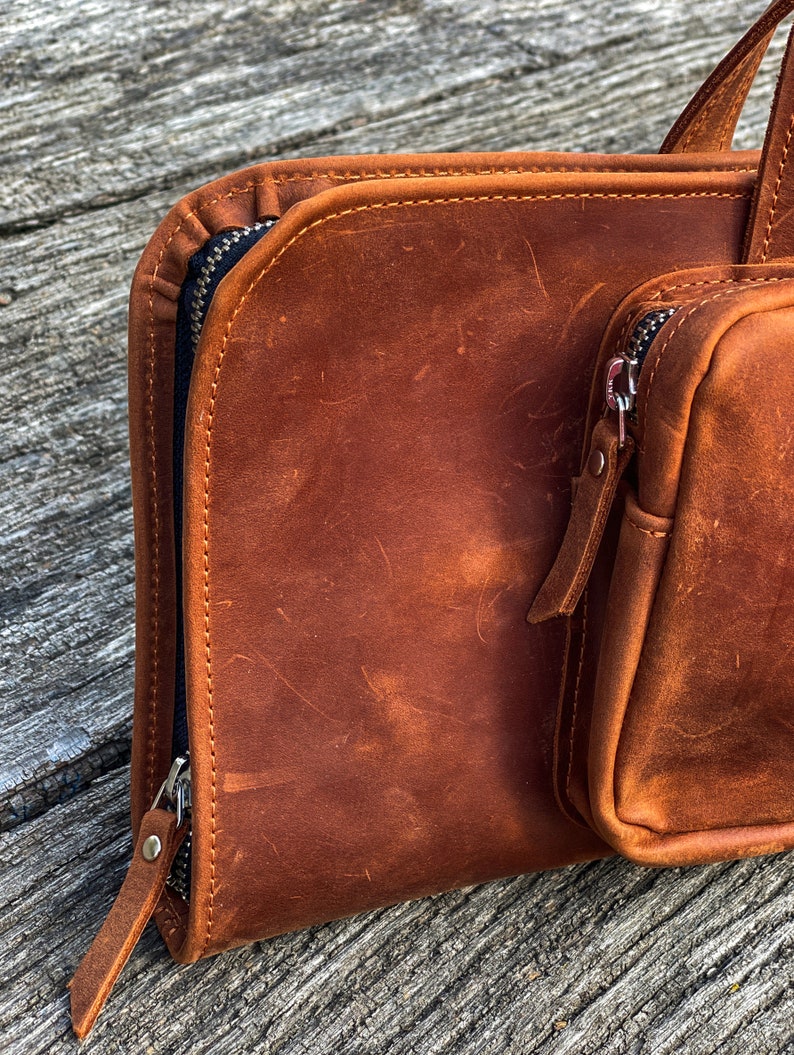 May include: A brown leather zippered pouch with a smaller zippered pouch attached. The main pouch has a flap that folds over and closes with a zipper. The smaller pouch is on the right side of the main pouch.