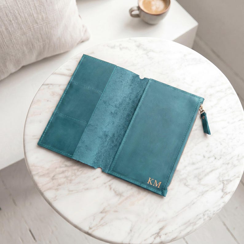May include: A teal leather wallet featuring a tri-fold design with multiple interior card slots and a zipped compartment. The exterior surface displays the embossed initials KM in the bottom right corner, complemented by a matching metal zip pull.
