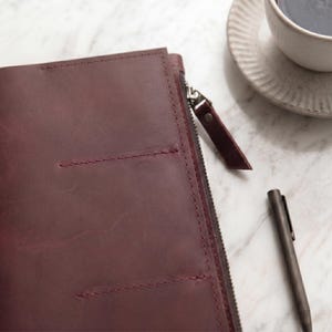 May include: A deep burgundy leather portfolio featuring a side zipper closure with a matching leather pull tab and two horizontal stitched pockets on the front cover. The portfolio is accompanied by a dark metallic pen and a ceramic cup.