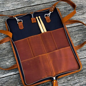 May include: A brown leather drumstick case with a zipper closure and two adjustable straps. The case has multiple pockets and is open to reveal two wooden drumsticks.