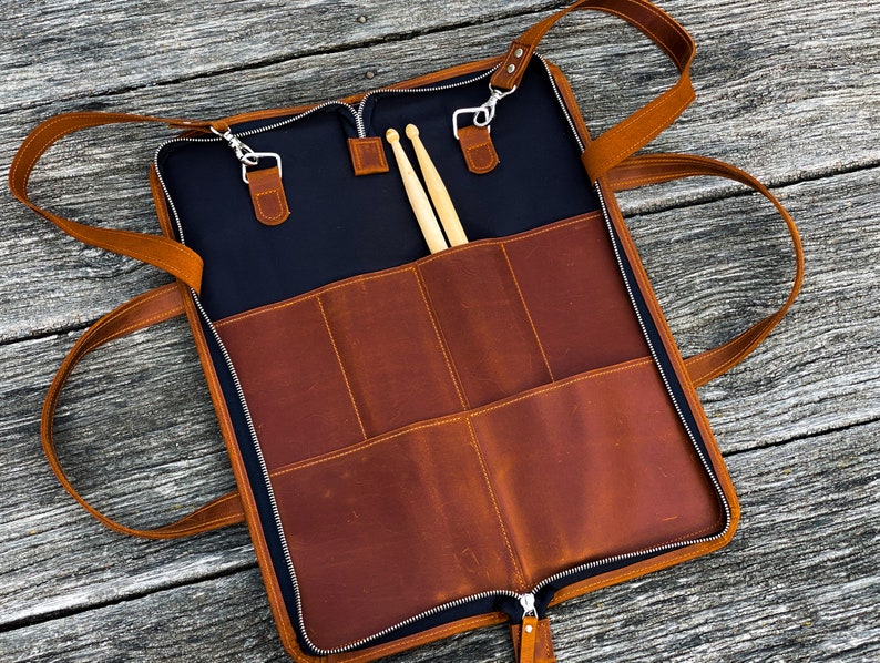 May include: A brown leather drumstick case with a zipper closure and two adjustable straps. The case has multiple pockets and is open to reveal two wooden drumsticks.