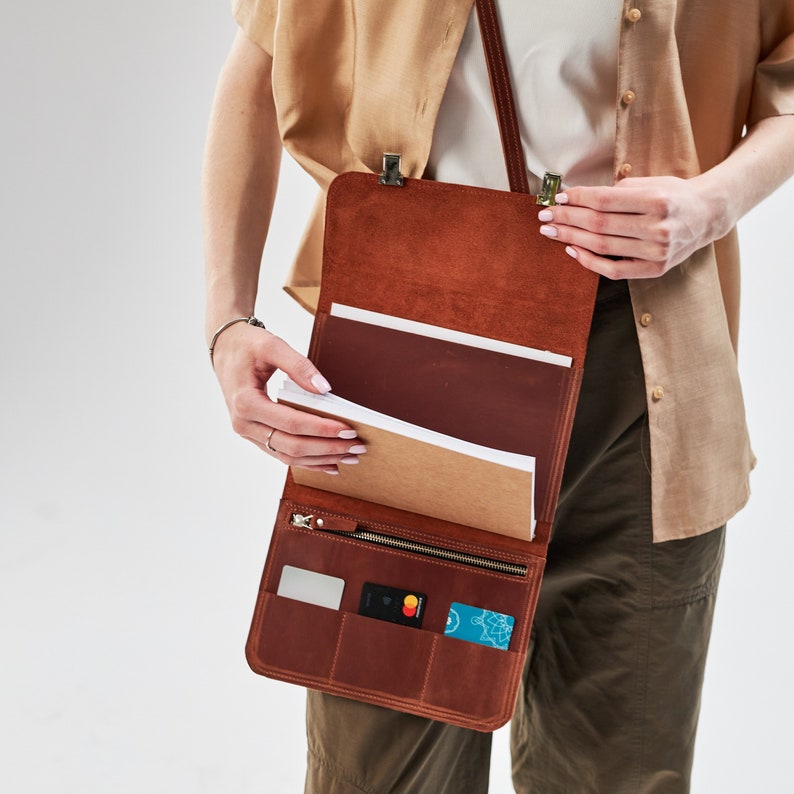 May include: A brown leather satchel bag with a zipper closure and multiple pockets. The bag is open and has a notebook and credit cards inside.