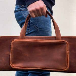 May include: A brown leather carrying case with a zippered pocket and two handles.