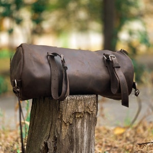 Puede incluir: Una bolsa de viaje de cuero marr&oacute;n con dos correas ajustables y hebillas plateadas. La bolsa est&aacute; sentada sobre un toc&oacute;n de &aacute;rbol de madera desgastado.