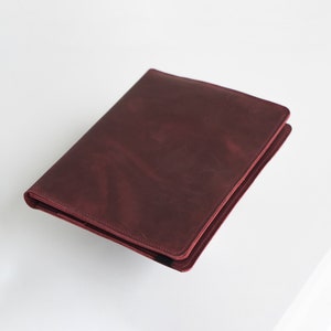 Puede incluir: Una carpeta de cuero burdeos con una superficie lisa y texturizada. La carpeta est&aacute; cerrada y tiene un brillo sutil.