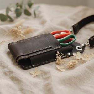 K&ouml;nnte beinhalten: Eine dunkelbraune Leder-Werkzeugtasche mit Schulterriemen. Die Tasche enth&auml;lt eine Schere mit roten und gr&uuml;nen Griffen. Das Leder ist mit einem helleren Faden gen&auml;ht. Der Hintergrund ist ein neutraler Stoff.