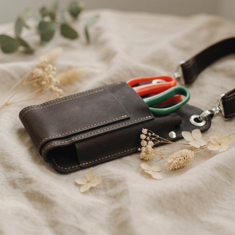 K&ouml;nnte beinhalten: Eine dunkelbraune Leder-Werkzeugtasche mit Schulterriemen. Die Tasche enth&auml;lt eine Schere mit roten und gr&uuml;nen Griffen. Das Leder ist mit einem helleren Faden gen&auml;ht. Der Hintergrund ist ein neutraler Stoff.