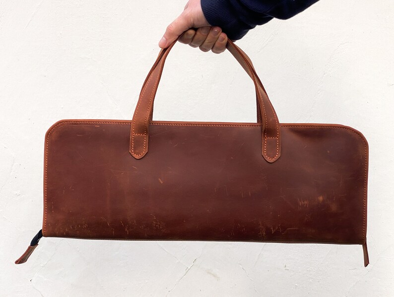 May include: A brown leather carrying case with two handles. The case has a zipper closure and is made of a distressed leather.