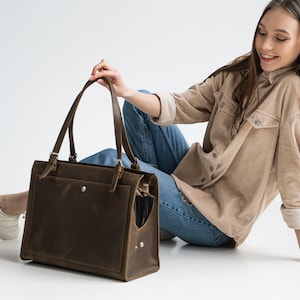 Pu&ograve; includere: Una borsa tote in pelle marrone con una tasca con cerniera e una chiusura a fibbia. La borsa &egrave; tenuta da una persona che indossa una camicia di velluto a coste marrone chiaro e jeans blu.