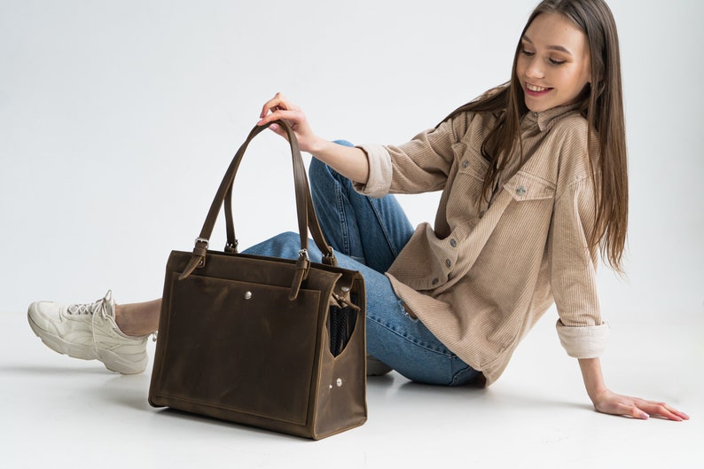 Pu&ograve; includere: Una borsa tote in pelle marrone con una tasca con cerniera e una chiusura a fibbia. La borsa &egrave; tenuta da una persona che indossa una camicia di velluto a coste marrone chiaro e jeans blu.