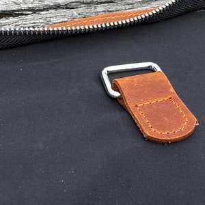 May include: Close-up of a brown leather strap with stitching detail attached to a silver metal buckle. The strap is attached to a black canvas bag.