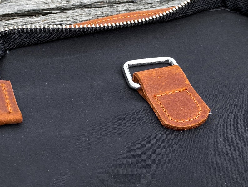 May include: Close-up of a brown leather strap with stitching detail attached to a silver metal buckle. The strap is attached to a black canvas bag.