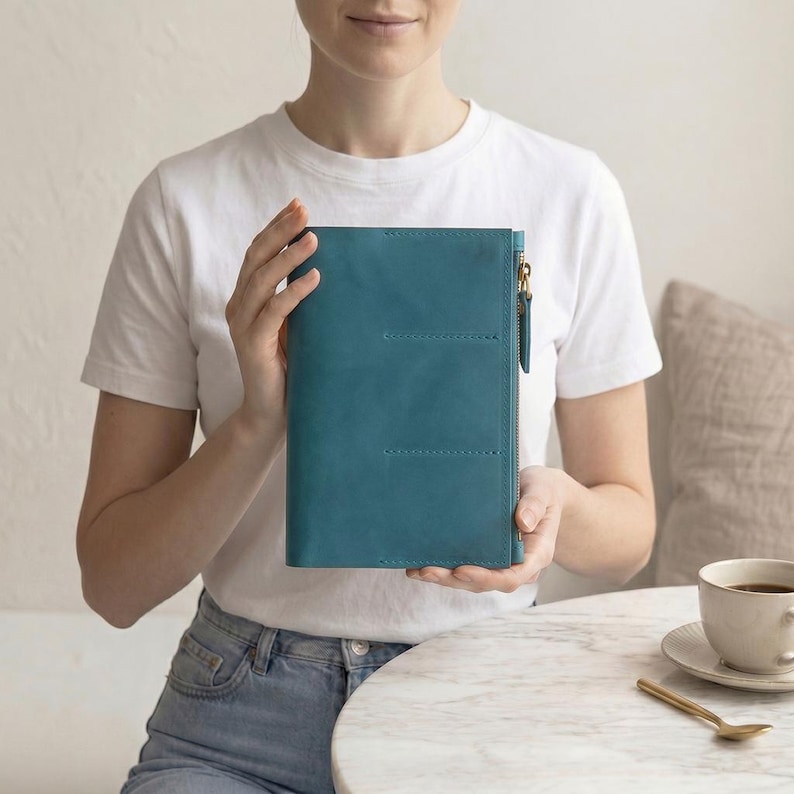 May include: A rectangular teal leather notebook cover featuring a vertical side zipper with a metallic pull. The cover has a smooth matte texture and includes two horizontal stitched lines on the front panel, providing a structured appearance.