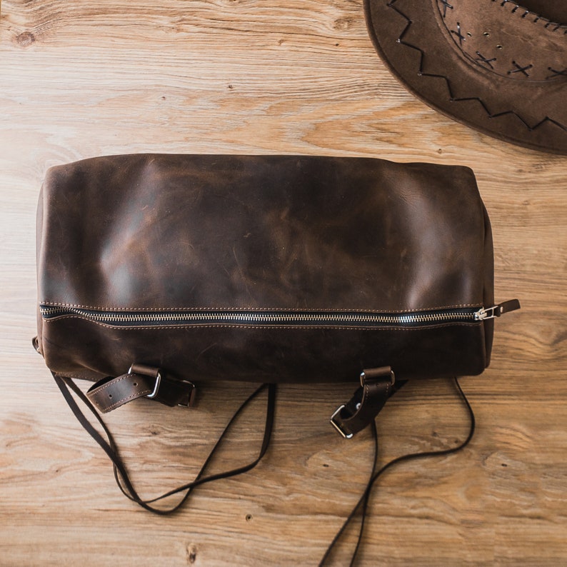 Puede incluir: Una bolsa de lona de cuero marr&oacute;n con cierre de cremallera y correas ajustables. La bolsa est&aacute; hecha de cuero desgastado y tiene un aspecto r&uacute;stico.