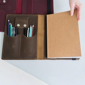 Puede incluir: Un portafolio de cuero marr&oacute;n con una solapa que se abre para revelar un cuaderno y m&uacute;ltiples ranuras para bol&iacute;grafos y l&aacute;pices.