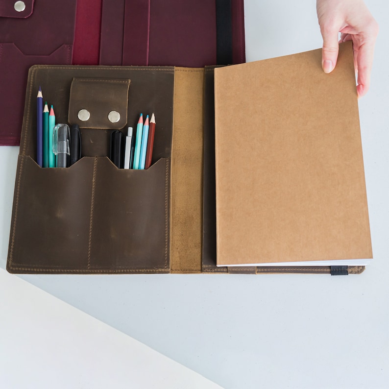 Puede incluir: Un portafolio de cuero marr&oacute;n con una solapa que se abre para revelar un cuaderno y m&uacute;ltiples ranuras para bol&iacute;grafos y l&aacute;pices.