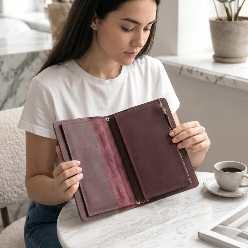 May include: A dark plum-colored leather organizer wallet featuring a tri-fold design, multiple interior card slots, and a vertical zippered pocket. The interior displays a smooth, matte finish with visible stitching along the edges and a metal zipper pull.