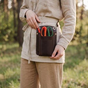 K&ouml;nnte beinhalten: Ein dunkelbrauner Leder-Werkzeugg&uuml;rtel mit mehreren Taschen und einer Schlaufe. Er enth&auml;lt eine orangefarbene und gr&uuml;ne Schere sowie einen roten Stift. Die Person tr&auml;gt ein beiges Hemd und eine Hose.