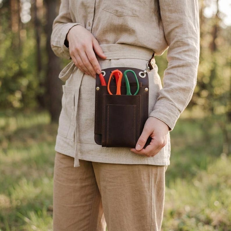 K&ouml;nnte beinhalten: Ein dunkelbrauner Leder-Werkzeugg&uuml;rtel mit mehreren Taschen und einer Schlaufe. Er enth&auml;lt eine orangefarbene und gr&uuml;ne Schere sowie einen roten Stift. Die Person tr&auml;gt ein beiges Hemd und eine Hose.